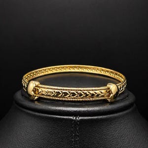 May include: A gold-coloured bangle bracelet with an intricate design. The bracelet features a central band with a repeating pattern and rounded end pieces. The bracelet is displayed on a black stand against a dark background.