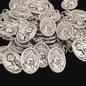 Puede incluir: Una colección de colgantes ovalados de metal plateado con un retrato en relieve de St. Carlo Acutis con el texto St. Carlo Acutis, o una representación de un cáliz con el texto JHS e Italy, cada uno con una pequeña anilla.