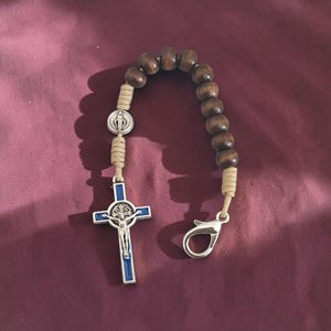 May include: A single-decade rosary featuring dark brown wooden beads strung on tan cord. It includes a silver-toned medal depicting a figure, a silver-toned crucifix with blue enamel inlay, and a silver-toned lobster claw clasp for attachment.