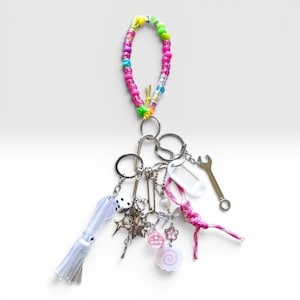 May include: A vibrant keychain with a beaded loop in pink, green, yellow, and blue. It includes charms such as a die, stars, a wrench, and a squid-shaped charm with a tassel. The background is plain white.