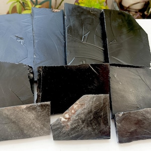 Könnte beinhalten: Eine Sammlung von neun rechteckigen Obsidianplatten mit einer glatten, reflektierenden schwarzen Oberfläche. Jedes Stück weist natürliche muschelige Bruchstellen und scharfe Kanten auf, wobei eine Platte einen kleinen hellbraunen Einschluss zeigt.