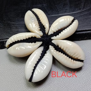 Op de afbeelding: Een bloemvormig ornament gemaakt van vijf witte schelpen, elk met een zwart midden. De schelpen zijn verbonden met zwart draad. Het woord "BLACK" is in het rood gedrukt.