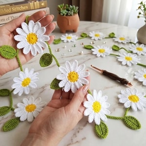 May include: Crocheted daisy chain garland with white petals, yellow centers, and green leaves. The handmade floral decor is displayed on a marble-like surface, with a crochet hook nearby. Perfect for spring or summer home decor.