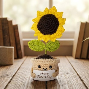 Op de afbeelding: Een gehaakte zonnebloem met een gele en bruine bloem, groene bladeren en een lachende pot met de tekst "Happy Father's Day". De handgemaakte decoratie staat op een houten oppervlak met een mok, boeken en een pot met varen op de achtergrond.
