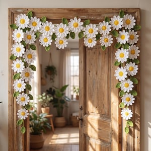 Puede incluir: Una guirnalda decorativa que presenta flores de margarita blancas tejidas a ganchillo con centros amarillos y hojas verdes, dispuestas para enmarcar un marco de puerta de madera. El arreglo floral consta de múltiples flores y follaje textil.