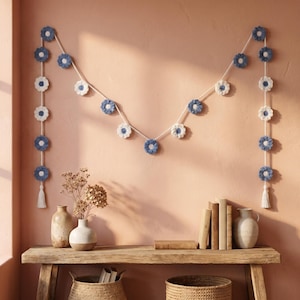 May include: A decorative garland featuring a series of crocheted flowers in alternating shades of dusty blue and cream, strung along a white cord. The garland hangs in a draped pattern with two vertical ends finished with small white yarn tassels.
