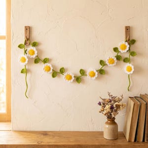 Puede incluir: Guirnalda de margaritas de ganchillo con pétalos blancos y centros amarillos, ensartada en vides verdes con hojas verdes. La guirnalda cuelga de una pared clara. Decoración del hogar.