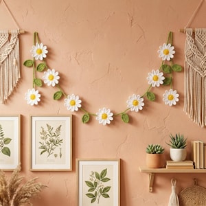 May include: A crocheted daisy garland with white petals and yellow centers, strung on a green vine with green leaves. The garland is hanging on a peach-colored wall, with framed botanical prints and decorative macrame.