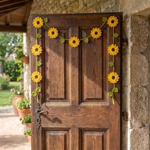 May include: A handmade crochet sunflower garland with yellow petals and brown centers, strung on green vines with green leaves. The garland is draped on a dark wooden door, creating a cheerful, rustic decoration.