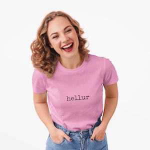 May include: A light pink short-sleeved t-shirt featuring the word "hellur" printed in a black, typewriter-style font across the chest. The garment has a classic crew neckline and is paired with blue denim jeans with visible metal buttons.