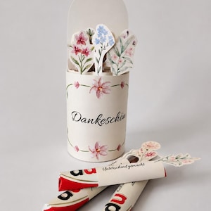 Puede incluir: Un recipiente cilíndrico de papel color crema decorado con ilustraciones florales y el texto Dankeschön. Dentro hay recortes de flores de papel. Tres barras de chocolate envueltas en papel blanco y rojo con el texto Unterschied gemacht.