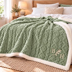May include: A sage green throw blanket with a textured cable knit pattern and a white sherpa fleece border. The blanket features an embroidered monogram with the letters B and H separated by a small leafy branch design in white thread.