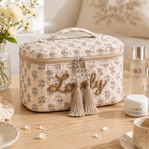 May include: A rectangular quilted vanity case in a cream fabric with a brown floral pattern. The front features the word Lovely embroidered in brown script, two silver zipper pulls with beige tassels, and a top handle for carrying.