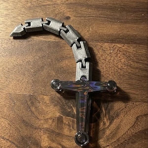 May include: A silver and black articulated fidget toy shaped like a sword. The handle has a cross-shaped design with a reflective, iridescent finish. The blade is made of connected, gray, rectangular links.