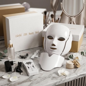 Könnte beinhalten: Ein weißes LED-Beauty-Maske mit passendem Nackenteil, Fernbedienung und Netzteil. Auf der Box steht "Colorful LED Beauty Mask". Dazu gehören eine kleine Flasche und eine Creme, die auf einer Marmoroberfläche angeordnet sind.
