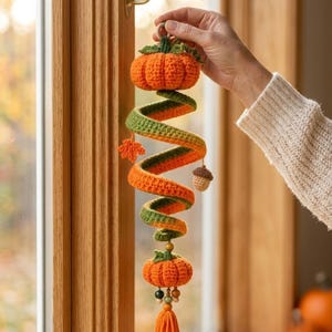 Könnte beinhalten: Eine hängende Häkeldekoration mit einem Spiraldesign aus orangefarbenem und grünem Garn. Das Stück enthält zwei kleine gehäkelte Kürbisse, ein winziges Ahornblatt und eine Eichel. Es ist mit Holzperlen und einer orangefarbenen Garnquaste versehen.