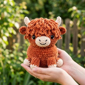 May include: A handmade, plush Highland cow toy. The cow is primarily brown with a curly texture, cream-colored horns, and a cream-colored face and feet. It has black eyes and a smiling expression. The toy is held in open hands against a blurred green background.