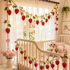 May include: A handmade crochet strawberry garland with white flowers and pink bows decorates a baby's crib. The garland is draped across the crib and window, creating a whimsical and charming nursery theme. The room also features a white crib, a vase of flowers, and a framed strawberry print.
