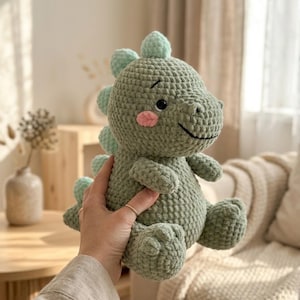 May include: A crocheted dinosaur toy in sage green yarn. The dinosaur has a smiling face with pink cheeks and mint green spikes. The soft toy is designed with a rounded shape and is suitable for children. The toy is made with a soft yarn.