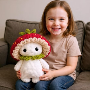 Puede incluir: Una muñeca de crochet con forma de seta con una gorra roja salpicada de puntos amarillos, un volante beige y un cuello verde. La muñeca tiene un cuerpo blanco, ojos negros y una pequeña sonrisa. La muñeca está siendo sostenida.