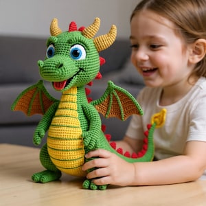Puede incluir: Un juguete de dragón tejido a ganchillo en verde y amarillo con ojos azules, alas naranjas y una lengua roja. El dragón tiene cuernos amarillos y una espina dorsal roja. El juguete está sobre una mesa de madera.