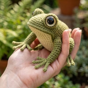 May include: A handmade crochet frog with large, expressive eyes. The frog is primarily green with a beige belly and detailed limbs. The amphibian is held in a hand, showcasing the craftsmanship of the amigurumi toy.