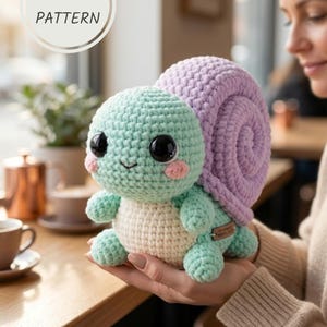 May include: A crocheted turtle plushie with a mint green head and limbs, a light purple shell, and a cream-colored body. The turtle has large black eyes and pink cheeks. The image includes the text "PDF PATTERN" in a white circle.
