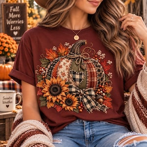 May include: A maroon t-shirt featuring a central graphic of a pumpkin decorated with various patterns including plaid, gingham, and leopard print. The pumpkin is surrounded by sunflowers, autumn leaves, berries, and a black and white checkered bow.