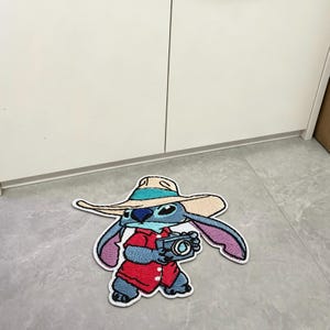 Puede incluir: Un parche bordado azul que muestra a un personaje con un sombrero beige de ala ancha con una banda turquesa, una camisa roja de botones y sosteniendo una c&aacute;mara. El personaje tiene orejas grandes, una nariz azul oscuro y pecho azul claro.