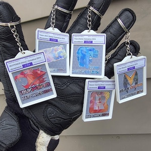 May include: A collection of keychains featuring various designs and text, including "iBUYPOWER Katowice 2014" and "Titan Katowice 2014". Each keychain has a silver chain and a white frame. The keychains are held in a black glove.