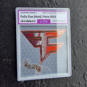 May include: A mint-graded sticker featuring the FaZe Clan logo in red and orange, with the text "FaZe Clan (Holo) | Paris 2023." The sticker is encased in a clear plastic holder. The bottom of the holder has a small gold-colored pin that says "Paris".