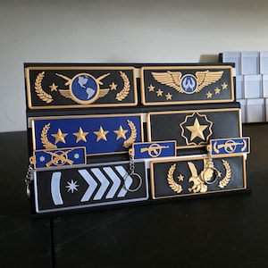 Könnte beinhalten: Ein schwarzer Präsentationsständer mit mehreren rechteckigen Schildern und Schlüsselanhängern, die goldene und blaue militärische Rangabzeichen zeigen. Die Motive umfassen Lorbeerkränze, Sterne, Flügel, einen Adler und Gewehre in einer gestuften Anordnung.