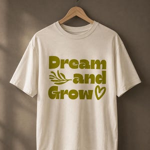 May include: Cream-colored t-shirt with the words "Dream and Grow" in a bold, olive green font. The design includes a sprig and a heart symbol. The shirt is hanging on a wooden hanger.