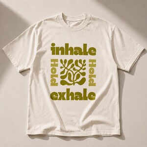 May include: Cream-colored t-shirt with a green design and text. The text reads "inhale, hold, exhale" with a green abstract floral design in the center. The shirt has short sleeves and a crew neck.