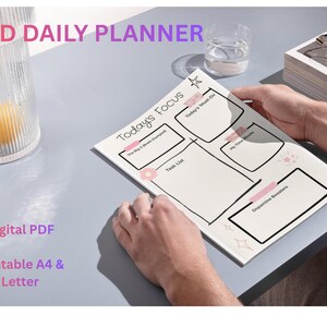 Könnte beinhalten: ADHS-Tagesplaner mit dem Text "Today's Focus" und Abschnitten für Aufgaben und Notizen. Der Planer wird von einer Person gehalten. Die Wörter "Digital PDF" und "Printable A4 & Letter" sind ebenfalls sichtbar.