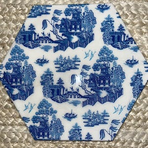Könnte beinhalten: Sechseckiger Keramikuntersetzer mit blau-weißem Chinoiserie-Design. Das Muster zeigt Pagoden, Brücken, Boote und Vögel. Der Untersetzer ist vor einem neutralen, gewebten Hintergrund platziert.