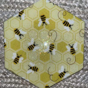 May include: Hexagonal coaster featuring a yellow honeycomb design and cartoon bees. The bees are black and yellow with white wings. The coaster has a glittery finish.