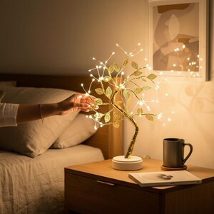 Puede incluir: Una lámpara decorativa dorada en forma de árbol con luces LED. La lámpara tiene una base blanca y está colocada sobre una mesita de noche de madera. Una taza gris y un libro también están en la mesita.
