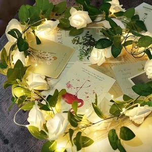 May include: A garland of white rose fairy lights with green leaves, illuminating vintage cards with botanical and flamingo illustrations. The lights create a warm glow, ideal for home decoration.