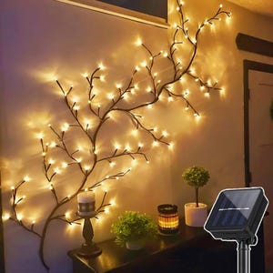 May include: A decorative wall-mounted branch with warm white LED lights. The branch is brown and has many small lights that resemble fireflies. A solar panel is visible in the lower right corner, along with a candle and small potted plants.