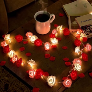 Puede incluir: Una guirnalda de luces en forma de corazón con rosas rojas y blancas. Una taza humeante y pétalos de rosa dispersos crean un ambiente romántico. También hay libros.