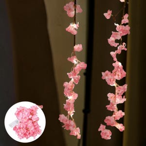 Pode incluir: Cordão de luzes de flores de cerejeira rosa. As luzes são enfiadas em um fio marrom e apresentam delicadas pétalas de flores rosa. As luzes são iluminadas, criando um brilho quente. Ideal para decoração.