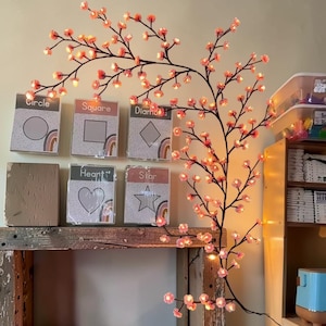 以下が含まれることがあります： ピンクの花と温白色のライトが付いた、装飾用の桜の枝。 枝の隣には、「円」、「正方形」、「ひし形」、「ハート」、「星」とラベル付けされた教育カードがあります。 カードには虹色のアクセントが付いています。