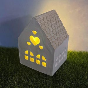 May include: A gray ceramic house-shaped candle holder with a textured roof and heart-shaped cutouts. The windows are illuminated with a warm yellow light, creating a cozy ambiance. The house sits on a bed of green grass.