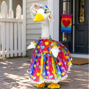 May include: A whimsical goose statue wearing a colorful dress adorned with heart shapes. The goose has a yellow beak and feet, and a white bonnet with lace trim. The dress features a rainbow of colors, making it a vibrant garden decoration.