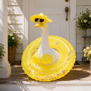 May include: A white goose figurine wearing a yellow polka dot sun hat, heart-shaped sunglasses, and a matching yellow polka dot bikini top, positioned inside a large, transparent yellow inflatable pool float filled with colorful confetti.