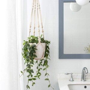May include: A hanging macrame plant holder with a white ceramic pot filled with a green leafy plant. The macrame is beige with wooden beads. The plant has long, trailing vines. The background includes a bathroom mirror and sink.