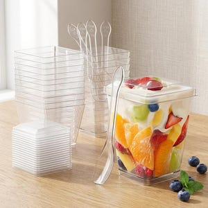 May include: A set of clear, square plastic dessert cups and matching transparent spoons. One cup is filled with a layered fruit salad containing orange slices, strawberries, blueberries, and grapes topped with a creamy white yogurt base.