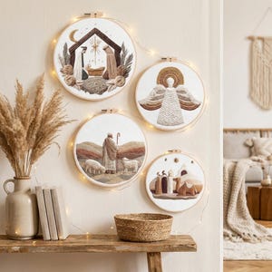 May include: Four circular embroidery hoops featuring religious scenes on white fabric. One depicts a nativity scene, another an angel, one a shepherd with sheep, and the last the three wise men with a camel. The hoops are illuminated by string lights.