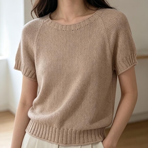 Può includere: Un top in maglia beige a maniche corte, con scollo tondo e orlo a coste. Il top ha una vestibilità ampia e maniche raglan. Il tessuto a maglia appare morbido e leggero, adatto alle temperature più calde. Lo stile generale è casual e confortevole.